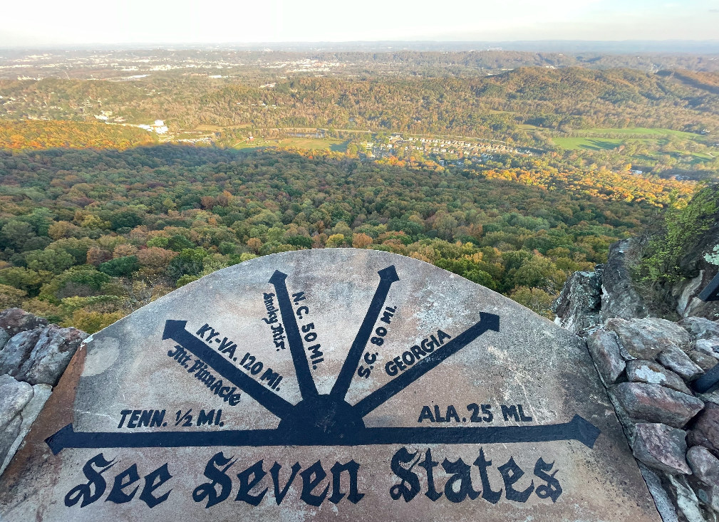 The See Seven States plaque atop Lover’s Leap: a stone marker painted with black arrows fanning out toward each claimed state, lettered Tenn. ½ mi., Mt. Pinnacle Ky.-Va. 120 mi., Smoky Mts. N.C. 50 mi., S.C. 80 mi., Georgia, and Ala. 25 mi., overlooking a forested valley.